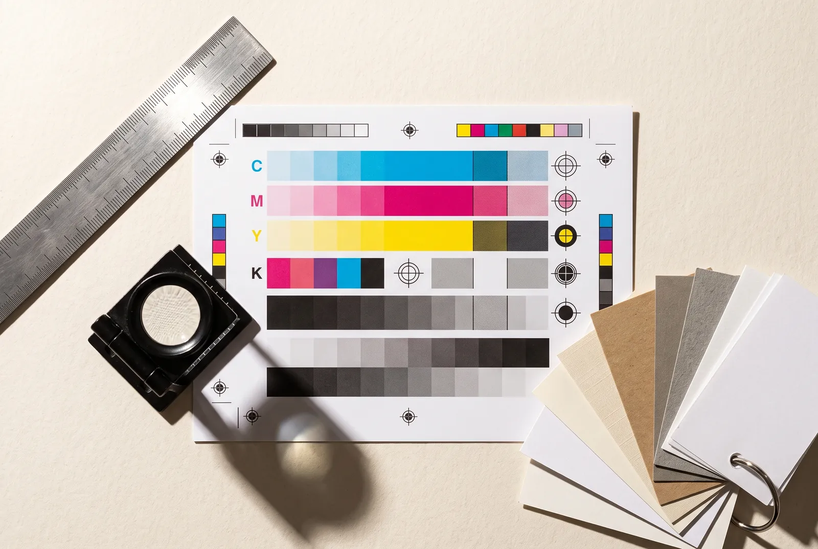 CMYK color calibration chart, steel ruler, loupe and paper stock swatches on a workbench.