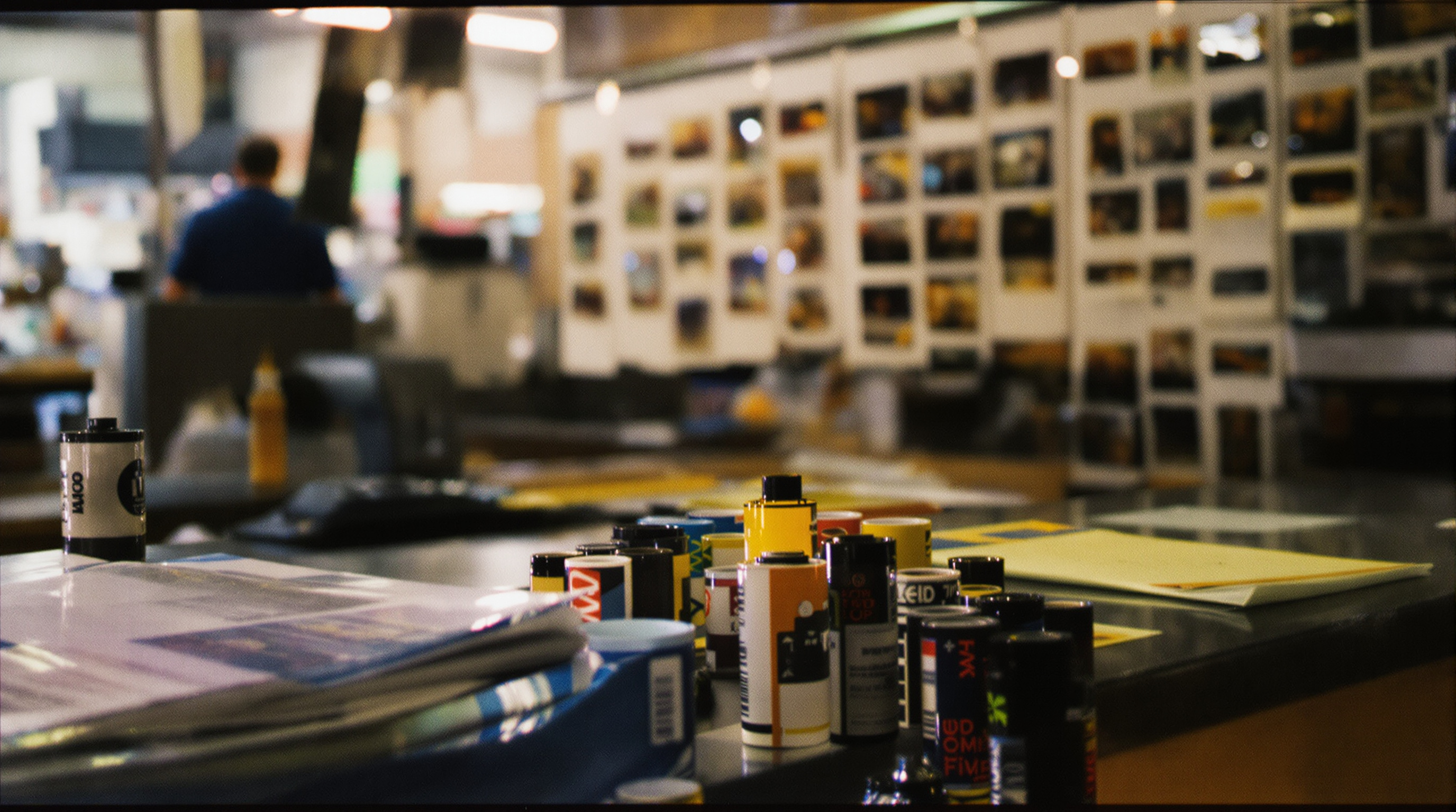 A warmly lit local film lab counter in Louisville, Kentucky, with rolls of 35mm color film, a film canister being opened, and developed color negatives hanging to dry in the background. Analog photography aesthetic, warm tones, shallow depth of field.