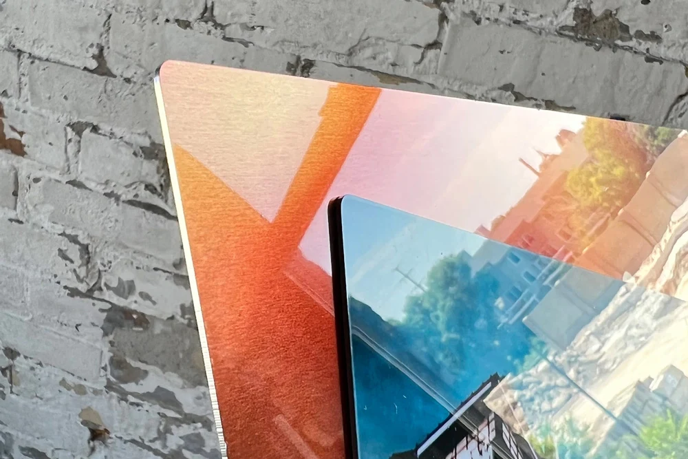 A close-up of glossy metal photo prints with vibrant colors and sharp details, stacked against a textured brick wall