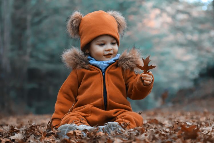 Toddler in orange coat and hat holding a fall leaf while sitting on ground