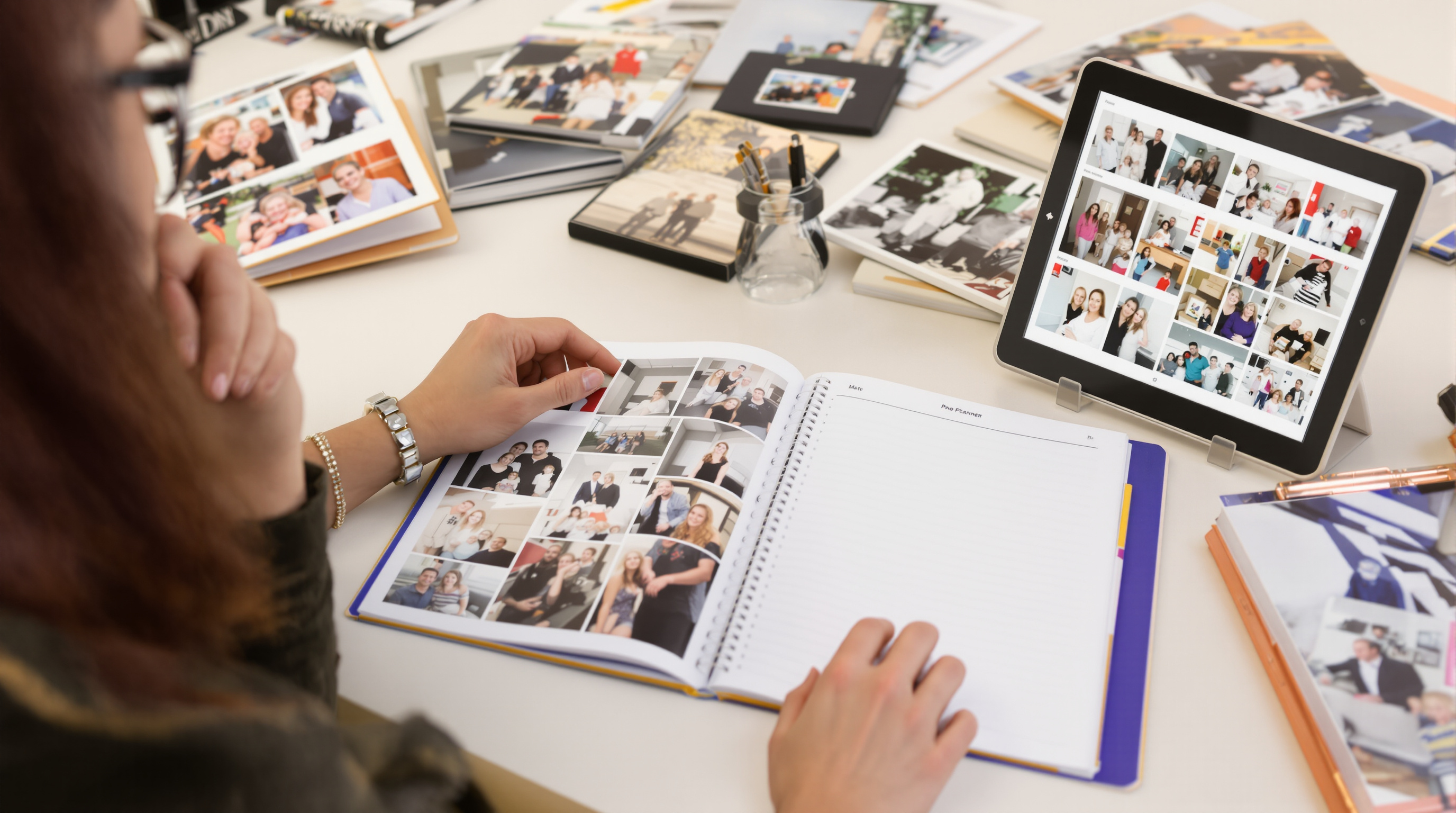 Person at desk organizing family photographs with photo albums, planner, and tablet displaying photo collections