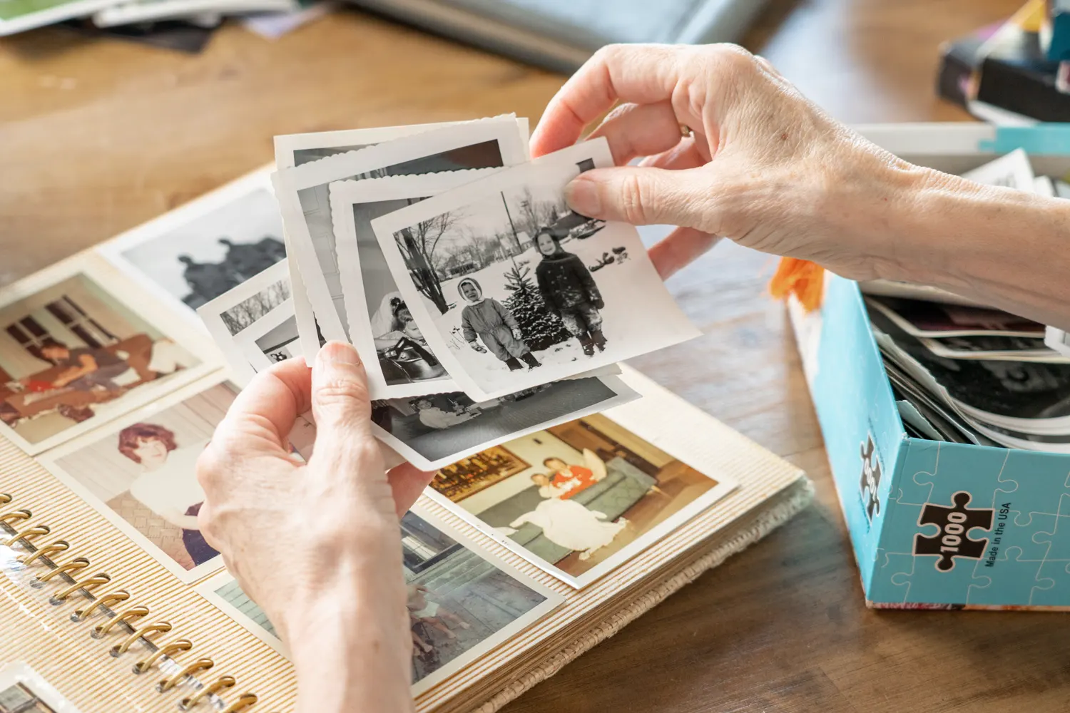 Hands holding vintage black-and-white photos above an open photo album and a box of pictures on a wooden table