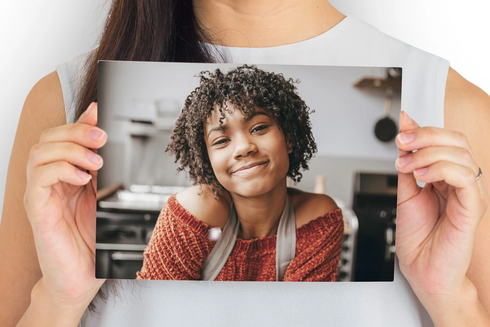 A person holding a cardstock photo print featuring a smiling young woman in a cozy sweater and apron, with a blurred kitchen background