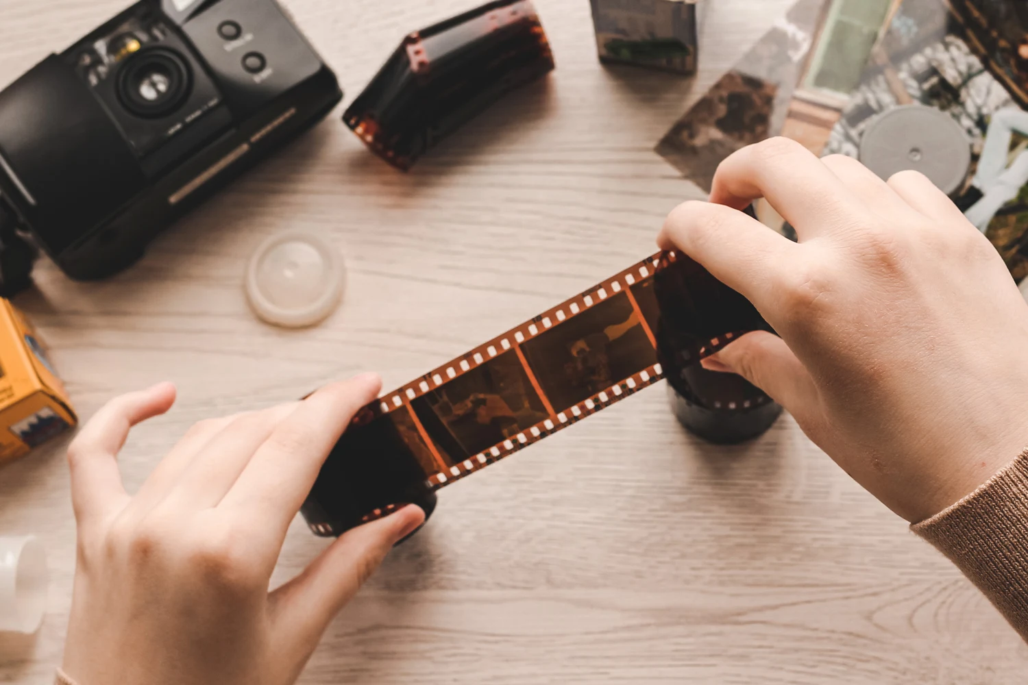 Hands holding 35mm film negatives over a table with a camera, film canisters, and photos, symbolizing film processing