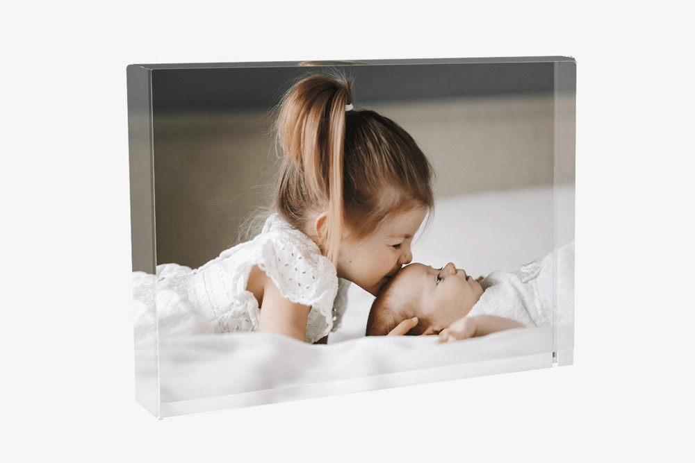 A custom glass photo print featuring a young girl kissing her baby sibling on the forehead, both dressed in white, lying on a bed
