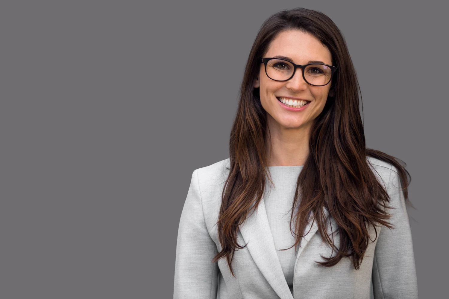 Professional headshot of a smiling woman with long brown hair, wearing glasses and a light gray blazer, on a gray background