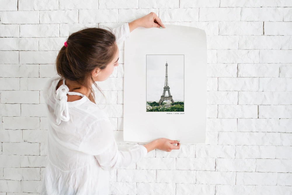A woman in a white blouse hangs a minimalist Eiffel Tower print on a white brick wall