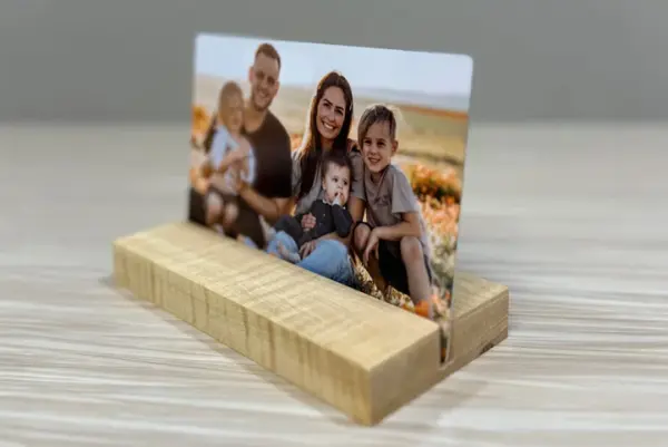 A custom metal photo print featuring a smiling family in a field of flowers, displayed in a wooden stand on a light wood surface