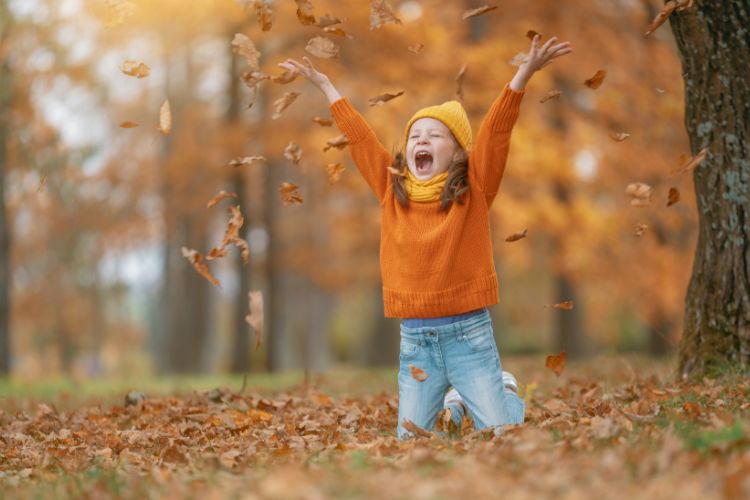Young girl in orange sweater throwing autumn leaves in the air