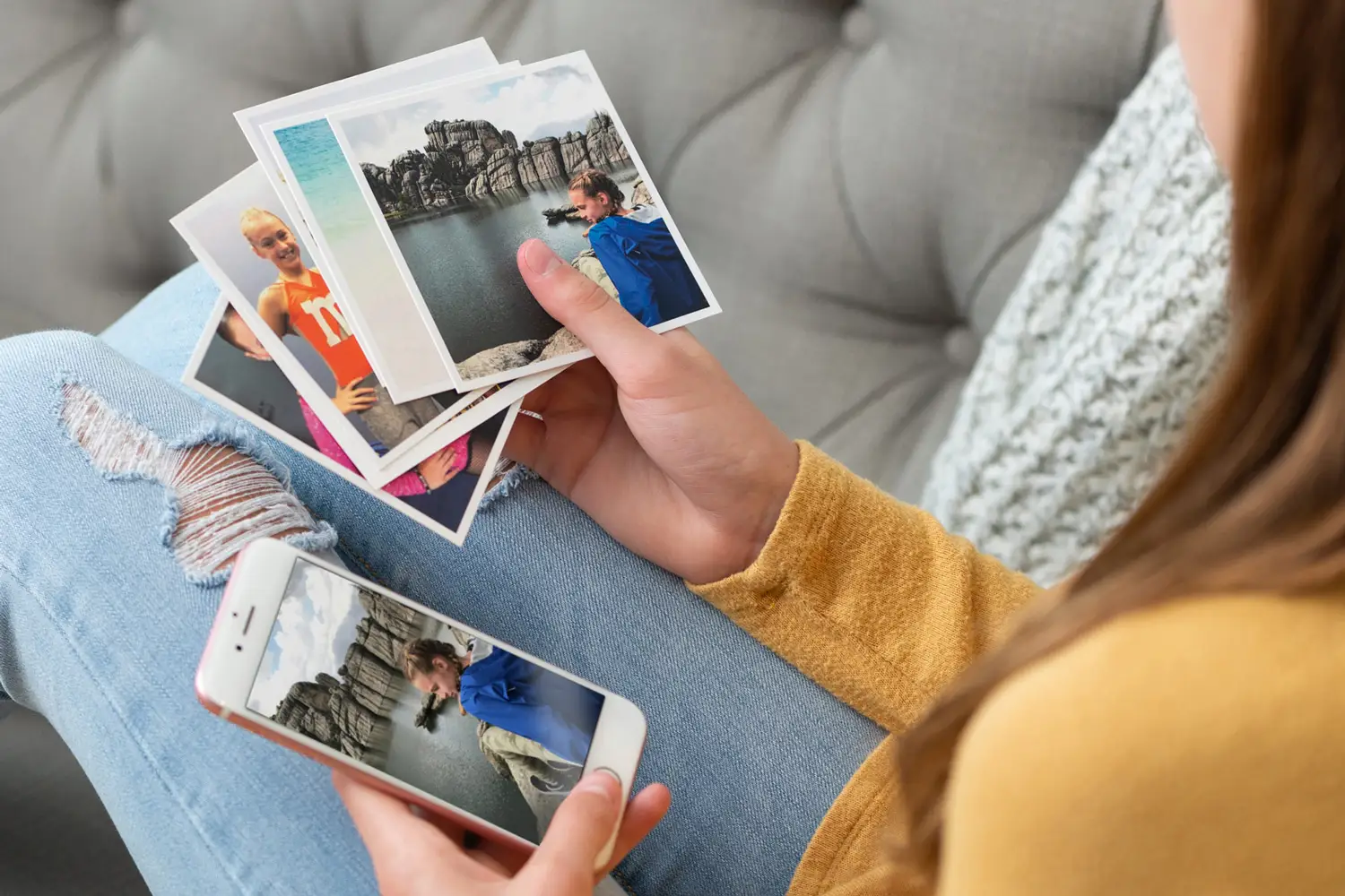 A teenage girl sitting on a gray couch holding printed photos in one hand and a smartphone displaying the same image in the other