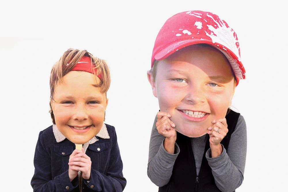 Two people holding oversized custom face cutouts in front of their faces, featuring smiling children, against a plain white background