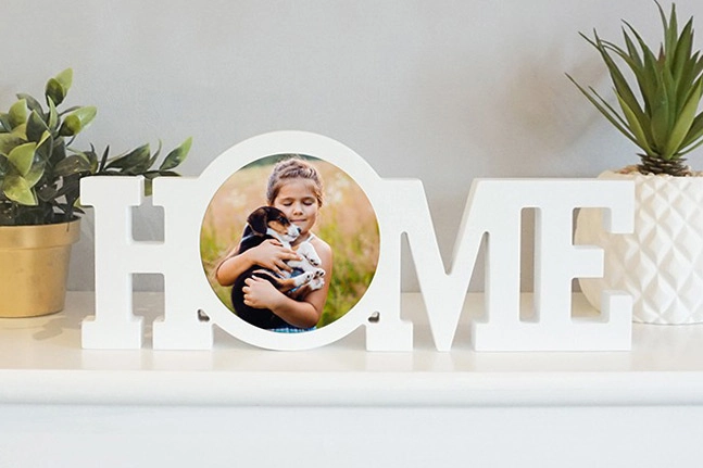 A white wooden HOME word block photo frame featuring a circular picture of a young girl hugging a puppy, displayed on a shelf with plants