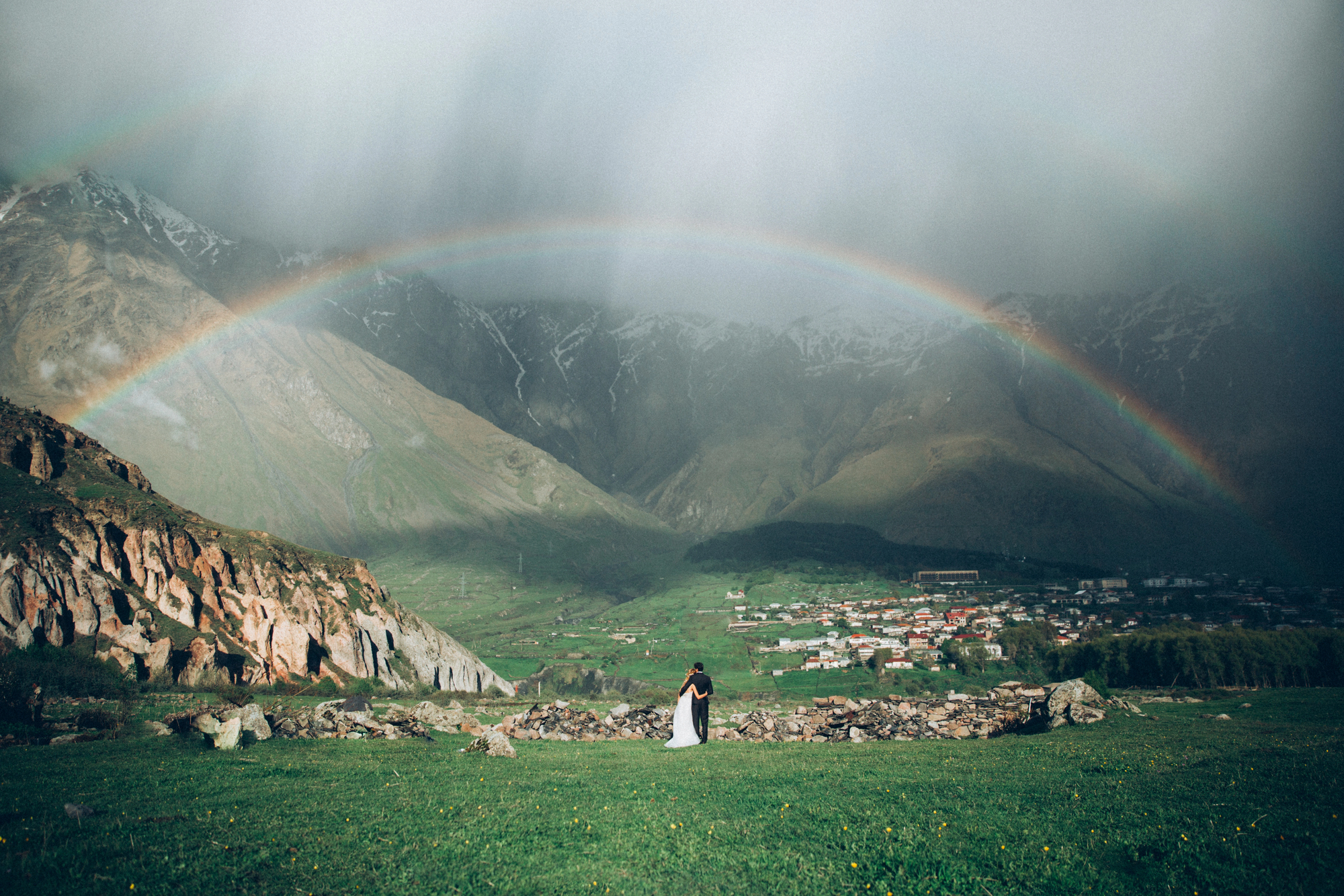 A couple on their wedding day standing under a rainbow on a mountain.