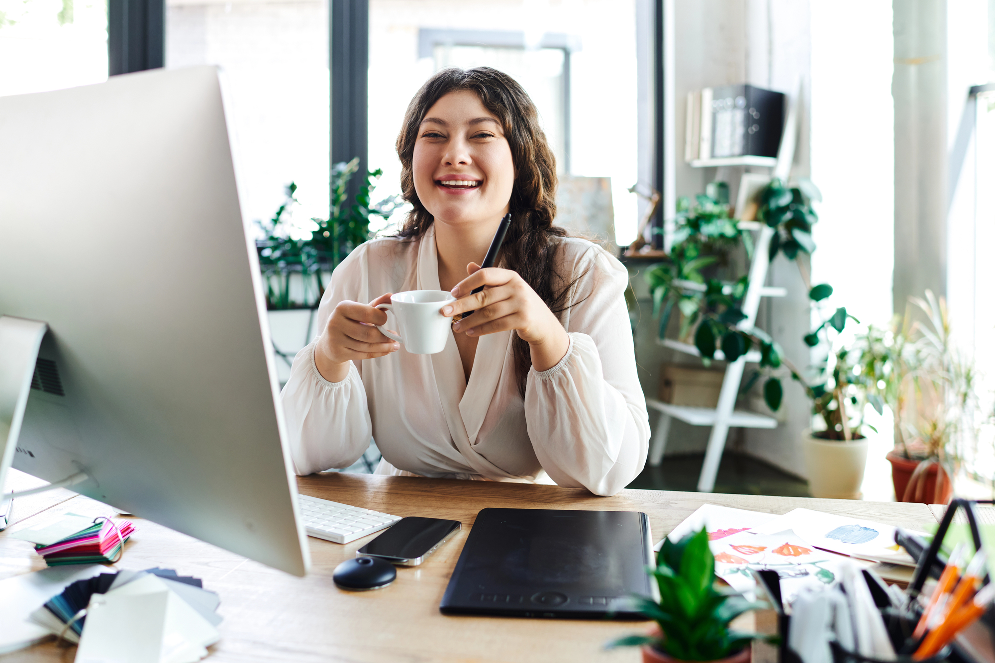 A person sits happily at their desk in their personalized workspace featuring plants, books, and an organized desktop. 