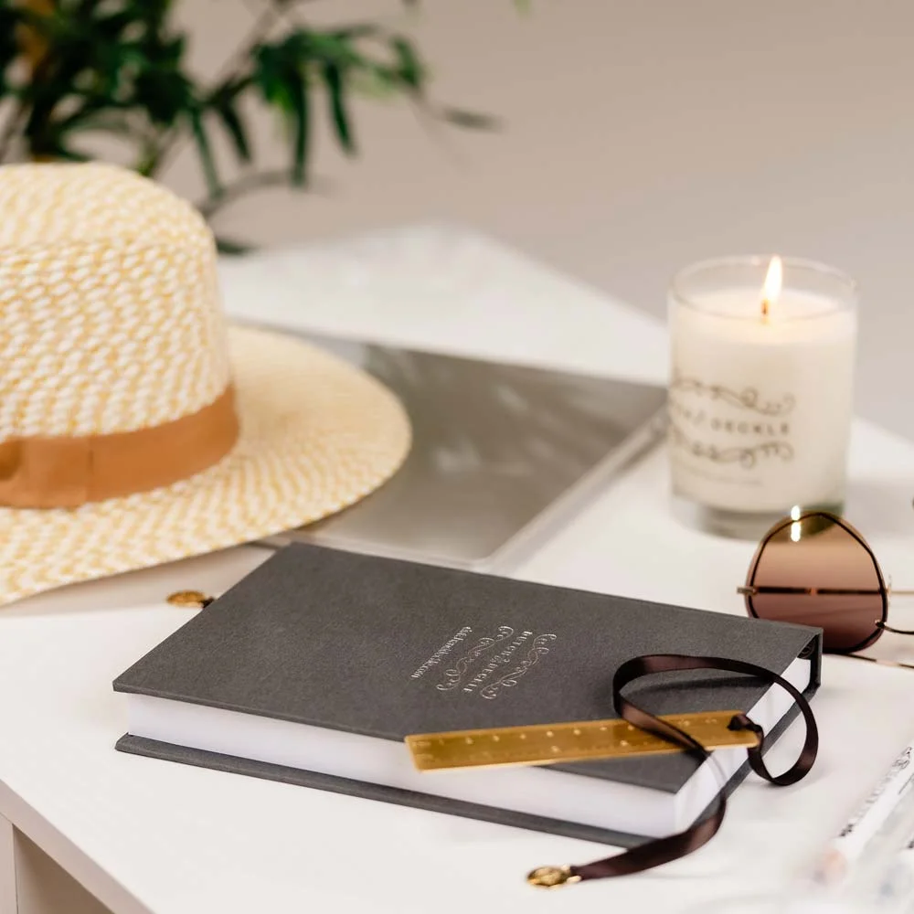 Tabletop with Dutch and Deckle candle, Vail dark grey linen journal, gold ruler bookmark and tan hat